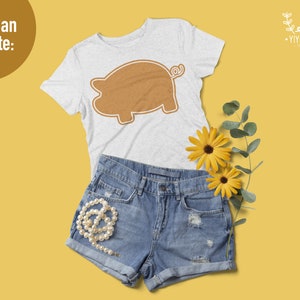 May include: A light gray short-sleeve t-shirt featuring a brown pig silhouette graphic, paired with blue denim shorts, a string of white pearls, and two yellow flowers. The text "You Can Create:" appears in the upper left corner of the image.