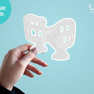 May include: A white sticker of a whimsical, multi-level house with a curved design. The sticker is being held by a hand against a light blue background.