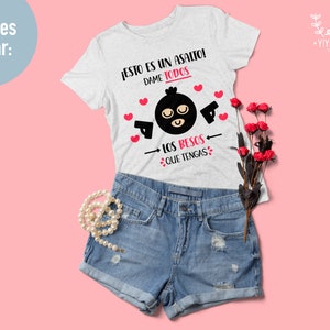 May include: A white t-shirt with a black and white illustration of a masked figure holding a gun. The text on the shirt reads "JESTO ES UN ASALTO! DAME TODOS LOS BESOS QUE TENGAS".