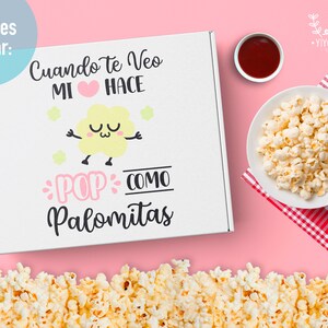 May include: A white box with the text "Cuando te veo mi coraz&oacute;n hace pop como palomitas" in black and pink lettering. The box has a cartoon popcorn with a face and stars around it. The box is on a pink background with a bowl of popcorn and a small bowl of red sauce.