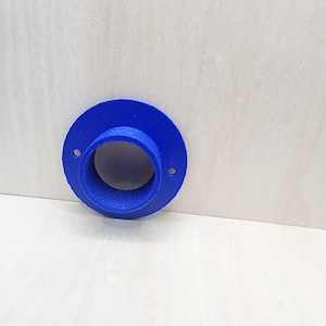 May include: A vibrant blue, 3D-printed flange with a circular design. The flange features a central opening and two small holes for mounting. The object is set against a light-colored wooden surface.