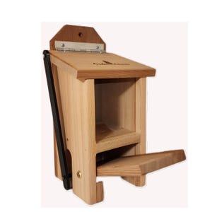 May include: A wooden birdhouse with a hinged roof and a small perch. The birdhouse is made of light-colored wood and has a rectangular shape with a small opening. A black metal arm supports the roof. The birdhouse has the text "Cuckoo Cabin" on the roof.