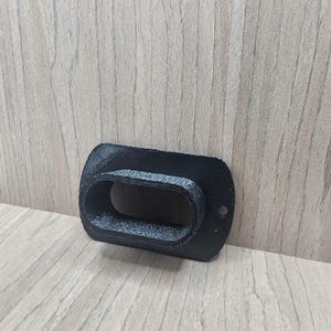 Puede incluir: Un objeto rectangular negro impreso en 3D con una abertura ovalada. El objeto tiene una superficie texturizada y un pequeño agujero en un lado. El artículo es probablemente un componente o parte de un producto más grande.