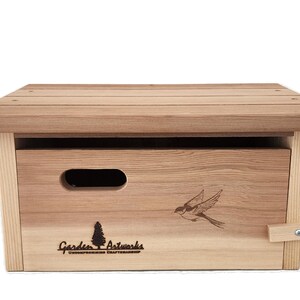 May include: A wooden birdhouse with a sloped roof and a rectangular body. The birdhouse features a cut-out handle, a bird graphic, and the text "Garden Artworks". The birdhouse is made of light brown wood.