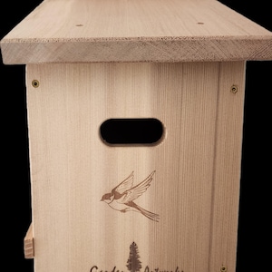 May include: A wooden birdhouse with a pitched roof and a small entrance hole. The front features a detailed engraving of a bird in flight and the text "Garden Artworks, Uncompromising Craftsmanship". The birdhouse is made of light-colored wood.