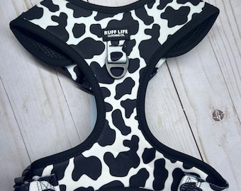 May include: Black and white cow print dog harness with a black adjustable strap and a silver metal D-ring. The harness has the text "RUFF LIFE CLOTHING CO." printed on it.