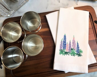 May include: Four silver shot glasses on a wooden cutting board with two white linen tea towels with embroidered purple and pink lupine flowers.