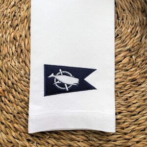 May include: White rectangular hand towel with a navy blue emblem featuring a whale, rope, and compass design. The towel has a textured surface and is displayed on a woven, brown surface.