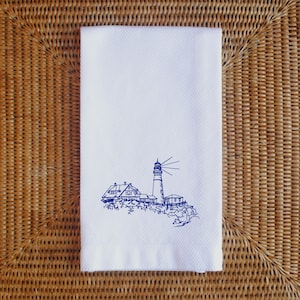May include: White hand towel with a textured surface and a blue line drawing of a lighthouse and buildings on a rocky coast. The lighthouse has a tall, striped tower with a light beam.