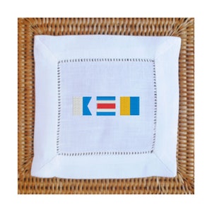 May include: White linen napkin with a nautical flag design embroidered in blue, red, yellow, and white. The flags spell out "KEI".