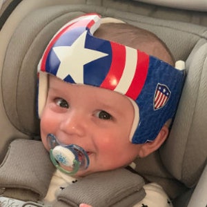 May include: A baby wearing a white, blue, and red helmet with a star and stripes design, reminiscent of a patriotic theme. The helmet has a white interior and is secured with straps. The baby is also wearing a pacifier.