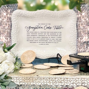 May include: A white ceramic serving tray with a scalloped edge and floral design. The tray features the text "The Origin of the Youngstown Cookie Table" and a description of the tradition. The tray is surrounded by several round, decorative cookies.