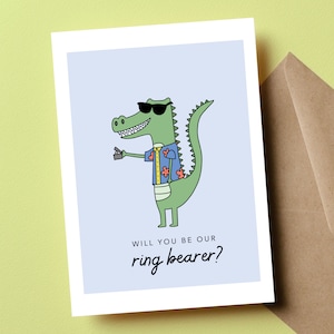 May include: A green alligator wearing sunglasses and a blue and pink Hawaiian shirt holds a small box in its hand. The text on the card reads "Will you be our ring bearer?"