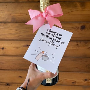 May include: A pink bow tied around the neck of a champagne bottle with a card attached that reads "Cheers to surviving the first year of parenthood" with a drawing of a baby bottle and a coffee cup.