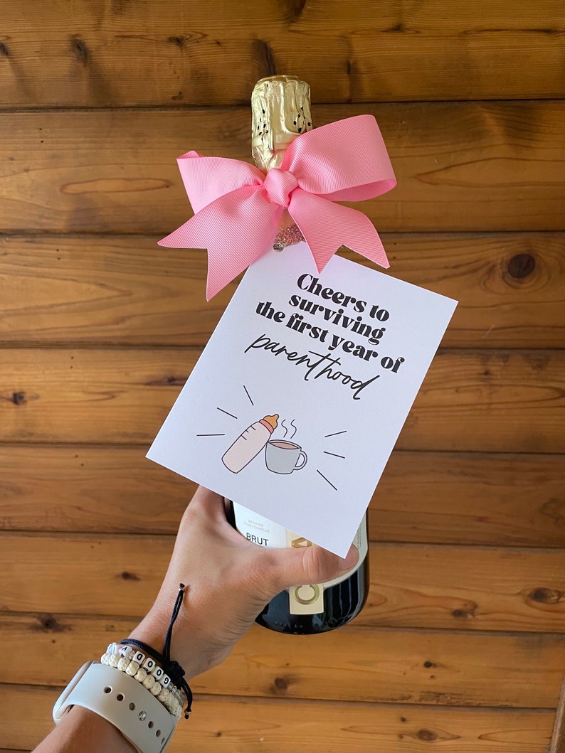 May include: A pink bow tied around the neck of a champagne bottle with a card attached that reads "Cheers to surviving the first year of parenthood" with a drawing of a baby bottle and a coffee cup.