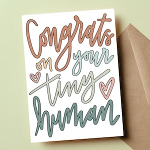 Könnte beinhalten: Eine weiße Karte mit dem Text "Congrats on your tiny human" in bunter Schrift. Der Text ist schwarz umrandet und hat Herzen drumherum.
