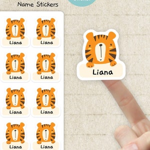 May include: A sheet of twenty rectangular waterproof stickers featuring a cartoon tiger face and the name Liana printed below. A single matching tiger sticker is shown separately. The text Little Tiger Name Stickers is visible at the top.