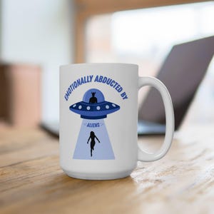 May include: White ceramic mug with a blue and black graphic of a UFO, an alien, and a person being abducted. The text on the mug reads "Emotionally Abducted by Aliens."