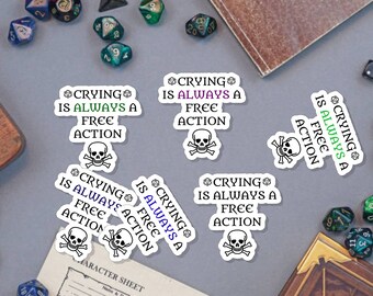May include: A set of six white stickers with a skull and crossbones graphic and the text "Crying is always a free action". The stickers are on a gray surface with dice and a book.