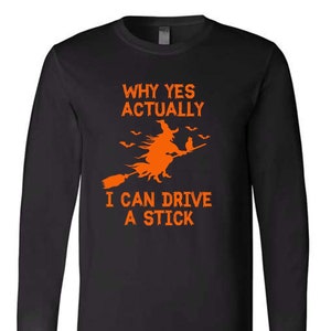 May include: Black long-sleeve t-shirt with an orange graphic of a witch flying on a broomstick with a cat. The text "Why yes actually I can drive a stick" is printed on the shirt.
