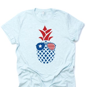May include: A light blue t-shirt with a pineapple wearing sunglasses with red, white, and blue stripes and a white star. The pineapple is a patriotic design.