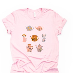May include: A pink t-shirt with a floral pattern of teapots and vases. The teapots and vases are decorated with flowers in shades of orange, yellow, and brown.