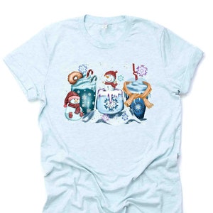May include: A light blue t-shirt with a winter-themed design featuring three snowmen holding mugs of hot chocolate with whipped cream and candy canes. The design is surrounded by snowflakes.