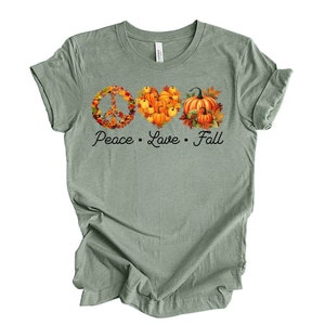 May include: A sage green t-shirt with a peace sign, heart, and pumpkin design made of fall leaves and pumpkins. The text "Peace Love Fall" is printed below the design.