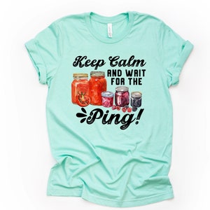 May include: A light teal t-shirt with the text "Keep Calm and Wait for the Ping!" above a watercolor illustration of several jars of preserves. The jars contain tomatoes, berries, and other fruits.