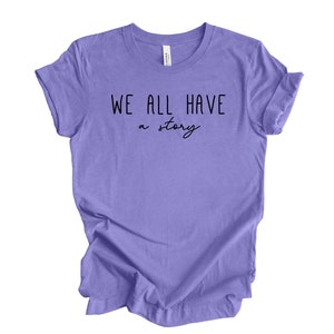 May include: Purple t-shirt with the text "WE ALL HAVE a story" in black.