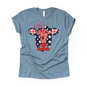 May include: A light blue t-shirt with a red, white, and blue patchwork cow face design. The cow has a red nose, white eyes, and a blue and red bow.