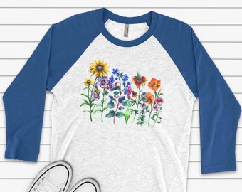 May include: A white and blue baseball tee shirt with a colorful floral print. The shirt has a blue raglan sleeve design.