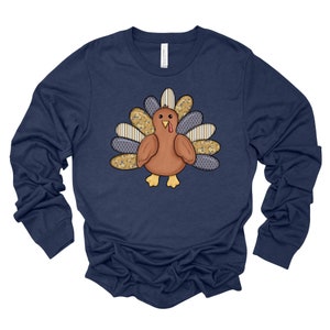 May include: A navy blue long-sleeved shirt with a patchwork turkey design. The turkey has a brown body, a red wattle, and a yellow beak. The patchwork design features blue, yellow, and white patterns.