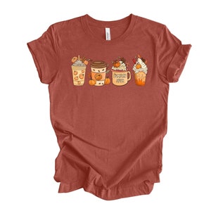 May include: A rust-colored t-shirt with a graphic of four fall-themed drinks. The drinks are a pumpkin spice latte, a pumpkin spice iced coffee, a pumpkin spice milkshake, and a pumpkin spice hot chocolate. The graphic is in a cartoon style and features pumpkins, leaves, and whipped cream.