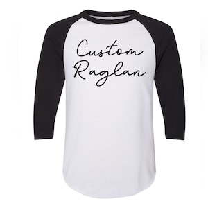 May include: White and black raglan shirt with three-quarter sleeves. The shirt features the text "Custom Raglan" in a cursive font across the front. The sleeves are black, and the body of the shirt is white.