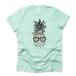 May include: A light green t-shirt featuring a graphic of a pineapple wearing a leopard print bandana and heart-shaped sunglasses. The pineapple has a black crown and is set against a white background. The shirt is made of a soft material.