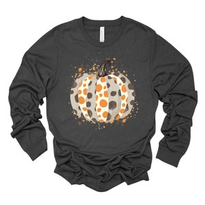 May include: Dark gray long-sleeve shirt featuring a watercolor-style pumpkin design. The pumpkin is white with orange and brown polka dots, and a brown stem. The design is surrounded by orange and brown paint splatters, perfect for fall.