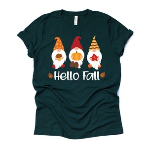 May include: A dark green t-shirt with a white graphic of three gnomes holding fall-themed items. The gnomes are wearing red, orange, and yellow hats. The text "Hello Fall" is written below the gnomes.
