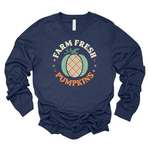May include: Navy blue long-sleeve shirt with a pumpkin graphic and the text "FARM FRESH PUMPKINS". The pumpkin design features a plaid center and floral accents, surrounded by stars.