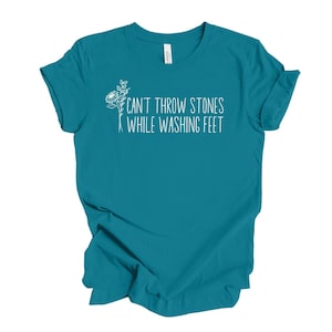 May include: Teal blue t-shirt with a white floral design and the text "Can't throw stones while washing feet."