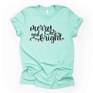 May include: A light blue t-shirt with the text "merry and bright" in black script. The text is surrounded by a string of colorful Christmas lights.