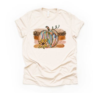 May include: A beige t-shirt with a colorful abstract pumpkin design. The pumpkin is surrounded by gold and brown brushstrokes and a small yellow flower. The design also includes a leopard print pattern.