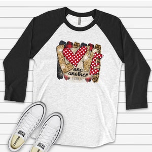 May include: A white and black baseball tee shirt with a glittery gold, red, and black graphic that says "Love one another 1 John 4:7".