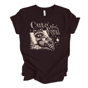 May include: A dark brown t-shirt with a graphic of a raccoon holding a cup of coffee and the text "Cute But Feral" in a vintage style font.