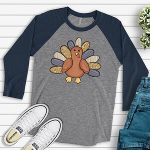 May include: A gray and navy blue baseball tee shirt with a patchwork turkey design. The turkey is brown with a red wattle and has a gray, white, and yellow patterned tail.
