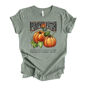 May include: Sage green t-shirt with a graphic featuring two orange pumpkins, green leaves, and the text "PUMPKIN PATCH EST. 1932." Additional text includes "HAYRIDER APPLE CIDER KETTLE CORN PUMPKIN PIE CARAMEL APPLES."
