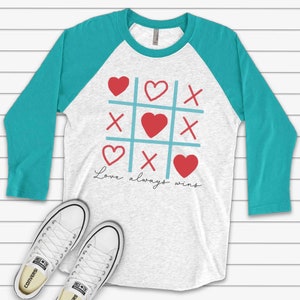 May include: A turquoise and white baseball tee shirt with a red heart tic-tac-toe design and the text "Love always wins".
