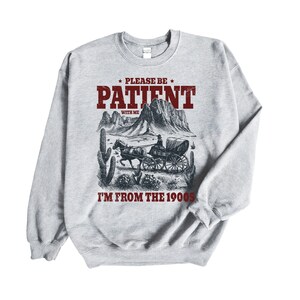 Könnte beinhalten: Grauer Pullover mit einem Vintage-Grafikdruck eines von Pferden gezogenen Wagens in einer Wüstenlandschaft. Der Text "Please be patient with me I'm from the 1900s" ist auf dem Pullover aufgedruckt.