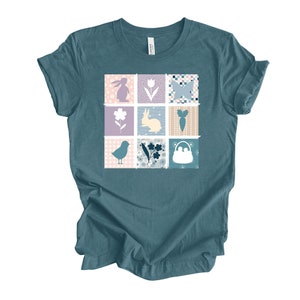 May include: Teal blue t-shirt with a patchwork design featuring a bunny, butterfly, flower, carrot, bird, and a basket. The design is in shades of pink, purple, blue, and white.