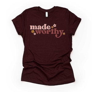 May include: A maroon t-shirt with the words "made worthy" in a retro font, accented with small floral graphics. The shirt is made of a soft material and has rolled-up sleeves.